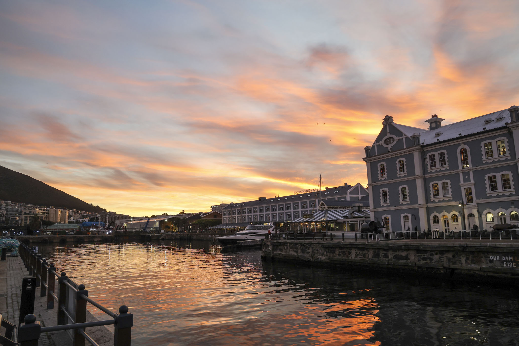 V&A Waterfront at sunset with historic buildings reflected in harbor water, orange and pink sky creating dramatic backdrop